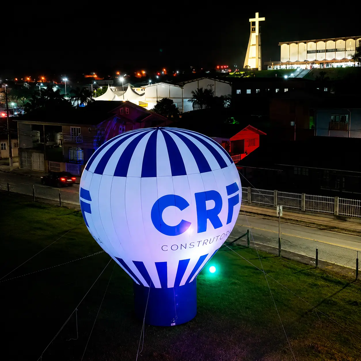 Vista aérea noturna do balão inflável gigante CRF Construtora de 10 metros brilhando com iluminação LED interna padrão da RVB Balões durante Festa dos Motoristas em Guabiruba SC, mostrando o impacto monumental visível a centenas de metros