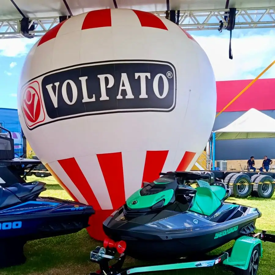 Balão Inflável Volpato — Feira Náutica Sea-Doo | RVB Balões Balão inflável vertical branco e vermelho com logotipo Volpato exposto em feira náutica ao lado de jet skis Sea-Doo, fabricado pela RVB Balões para revenda náutica em estande de exposição