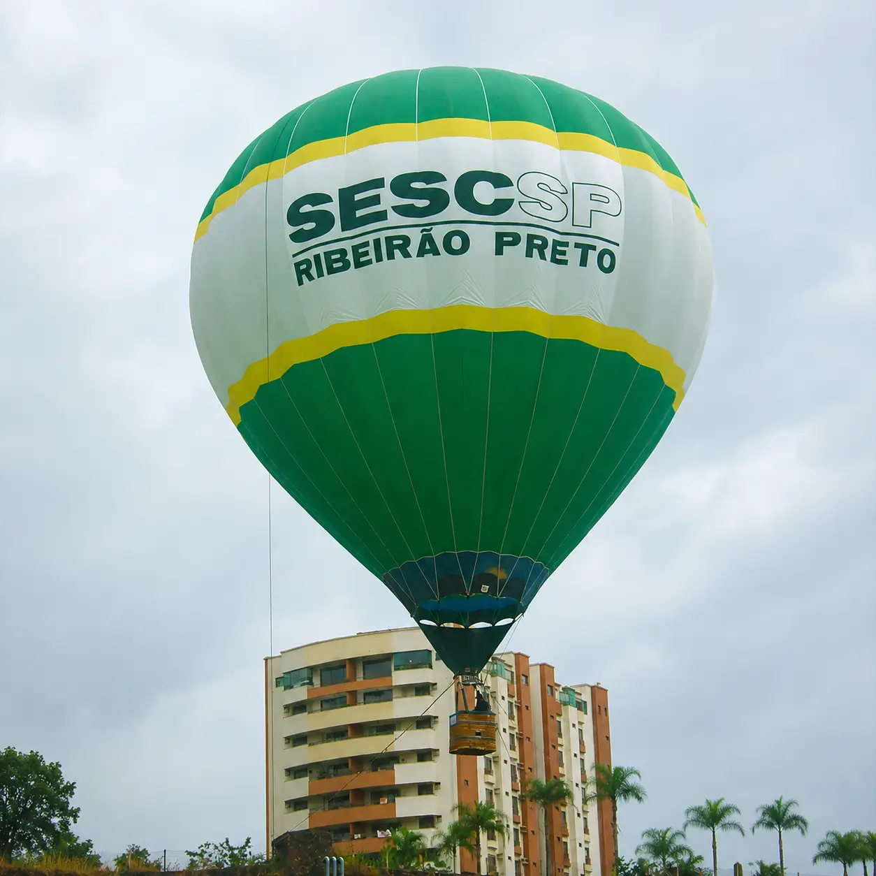Voo Cativo com Balão Personalizado SESC SP Ribeirão Preto — Ativação Institucional Urbana | RVB Balões Balão de ar quente personalizado SESC SP Ribeirão Preto em voo cativo sobre centro urbano com edifícios ao fundo, operação RVB Balões em ativação institucional