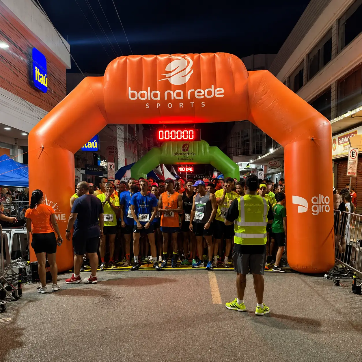 Portal Inflável Bola na Rede Sports — Largada de Corrida | RVB Balões Portal inflável laranja Bola na Rede Sports na largada de corrida de rua noturna com cronômetro digital e atletas alinhados, fabricado pela RVB Balões para evento esportivo com patrocínio Itaú