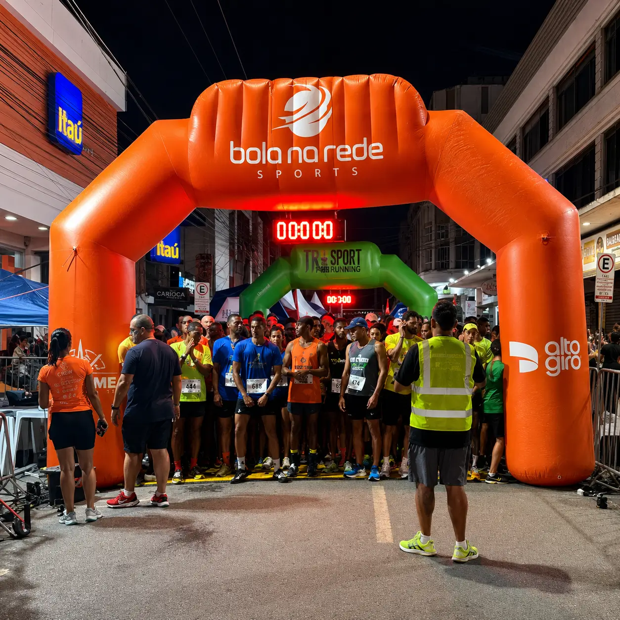 Portal inflável personalizado para corrida de rua durante evento esportivo noturno, com atletas posicionados na largada sob estrutura inflável promocional da Bola na Rede Sports.